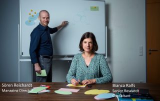 Sören Reichardt, Managing Director, at the whiteboard in the background, Focus on Bianca Ralfs, Senior Research Consultant, in the foreground with pen and sheets of paper