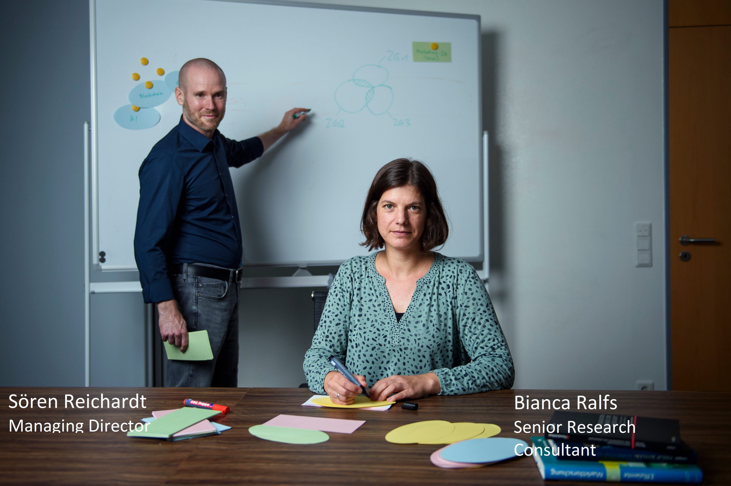 Sören Reichardt, Managing Director, at the whiteboard in the background, Focus on Bianca Ralfs, Senior Research Consultant, in the foreground with pen and sheets of paper